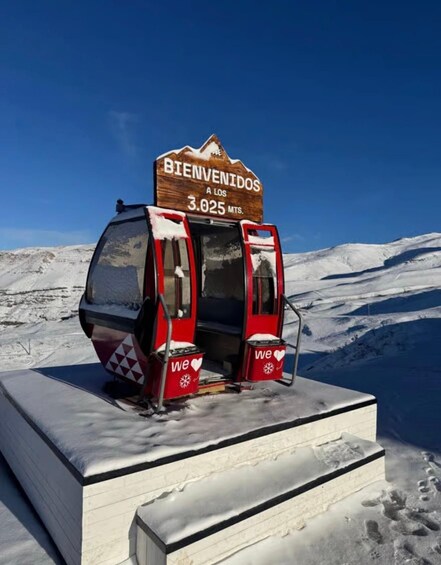 A red and white cable car with a wooden sign reading 'BIENVENIDOS A LOS 3.025 MTS.' in a snowy mountainous area. 3