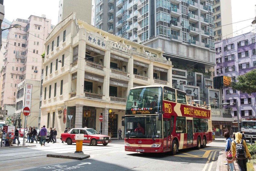 Punainen, kaksikerroksinen bussiyhtiön «BIGBUS HONG KONG» -merkinnällä varustettu bussi ajaa kaupungin kadulla, jossa on jalankulkijoita ja rakennuksia. 5