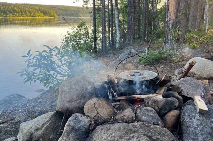 A campfire with a pot on a portable stove, surrounded by rocks and trees near a calm lake. 2