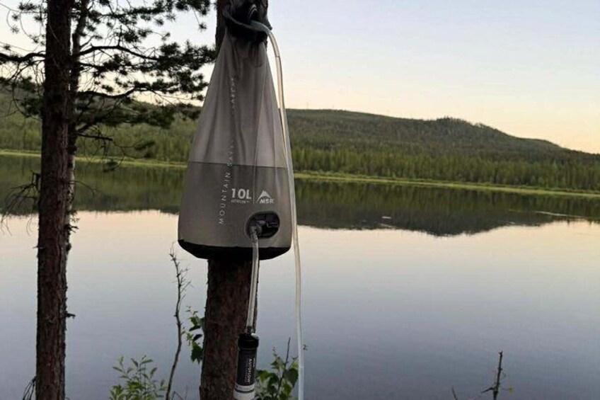 A portable water filter hanging on a tree by a calm lake with a forested hill in the background. 3