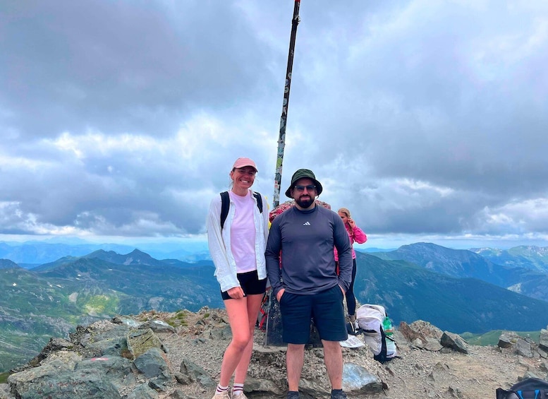 Two people standing on a rocky mountain peak with a cloudy sky and distant mountains in the background. 4