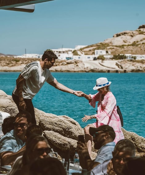 A man and woman shaking hands on a rocky seaside with a crowd in the foreground and buildings in the background. 5