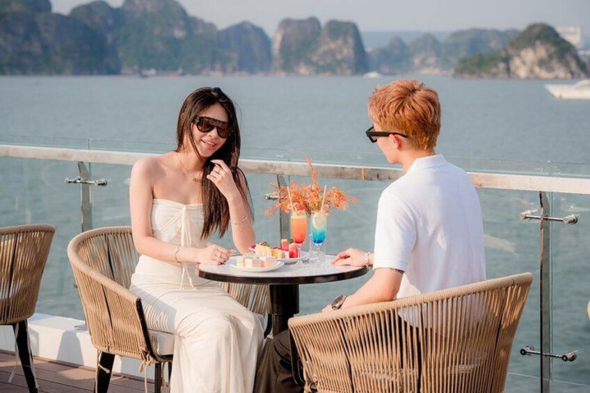 Two people sitting at a table with drinks and a dessert, overlooking a body of water with cliffs in the background. 5