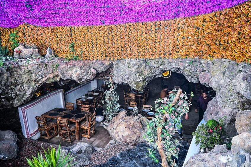 En hulelignende struktur med borde og stole indeni, omgivet af farverige blomster. 4
