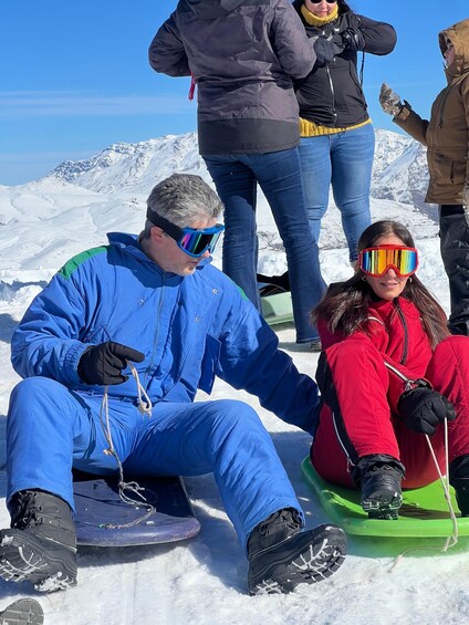 A person in blue snow pants and a blue jacket sitting on a green sled, another person in red snow pants and a red jacket sitting on a green sled, and a person in a brown jacket standing in the background. 4