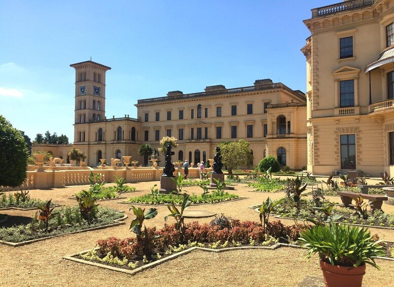 A historic courtyard with a clock tower, ornate buildings, and a well-maintained garden with statues and plants. 2