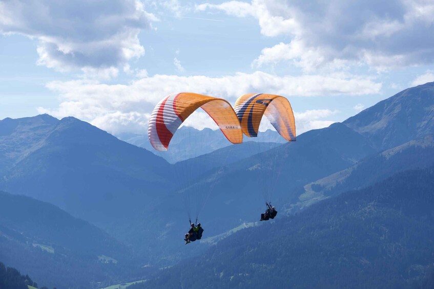 Two paragliders with orange and white canopies flying over a mountainous landscape. 1