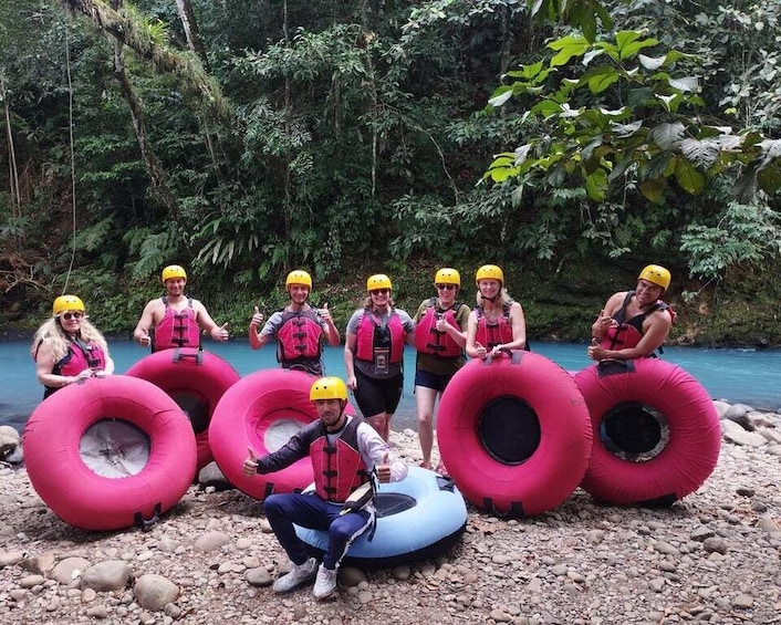 A group of people in life jackets and helmets, holding inflatable tubes on a rocky riverbank. 5