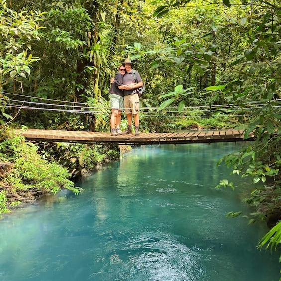 Two people standing on a wooden bridge over a clear blue river, surrounded by dense green foliage. 1