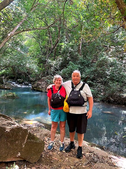 Two people standing by a clear stream in a forested area. 3