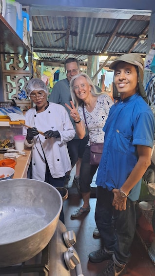 A group of people in a market setting with a large metal bowl and cooking utensils on the counter. 1