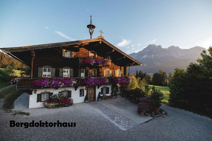 A traditional wooden house with a balcony, surrounded by a gravel driveway and flowering plants, set against a mountainous backdrop. 2