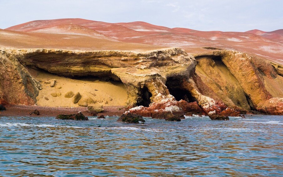 A coastal rock formation with a cave entrance, surrounded by water and red-hued hills. 4