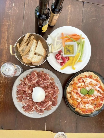 A table with a plate of prosciutto, a pizza, a colander of bread, a plate of vegetables, and two bottles of olive oil. 1