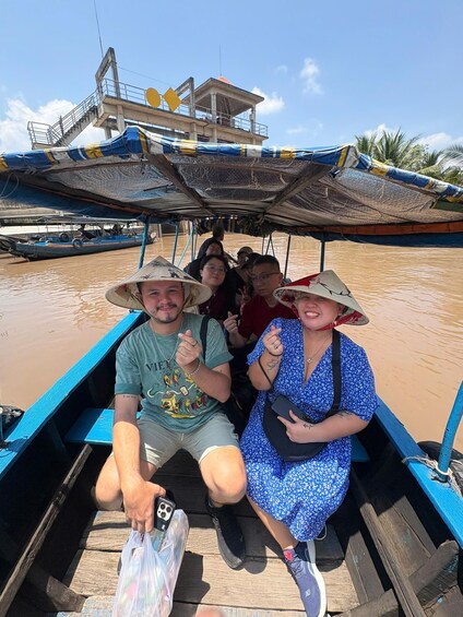 A group of people on a wooden boat with a blue canopy, floating on a river with a bridge in the background. 2