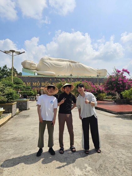 A large Buddha statue, three people in traditional attire, and a sign with foreign script. 3