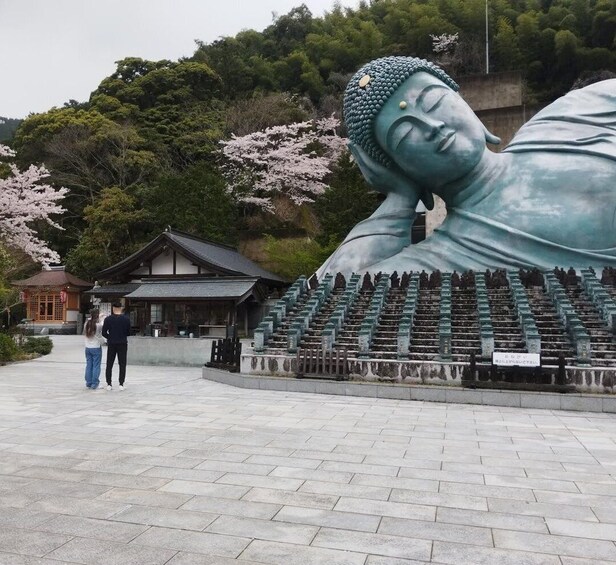 A large reclining Buddha statue with a series of steps leading up to it, surrounded by cherry blossoms and traditional architecture. 5