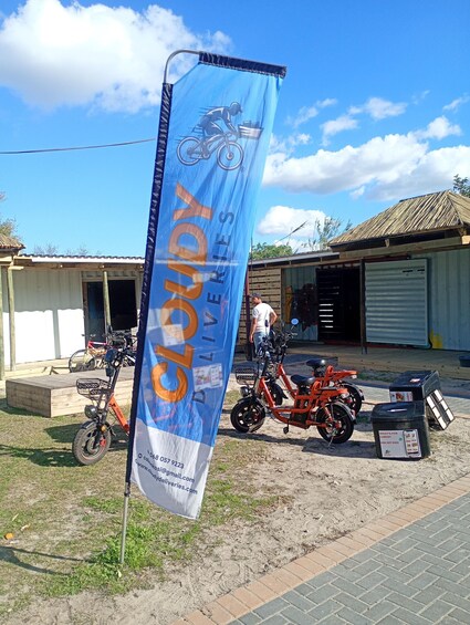 A blue banner with 'CLOTH DELIVERY' and contact information, a red scooter, and a building with a corrugated metal roof. 3