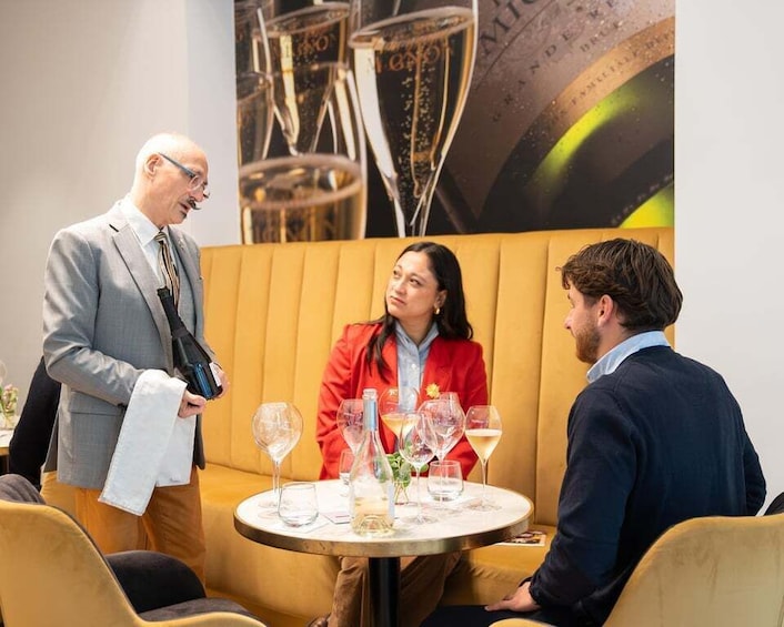 A group of people in a modern indoor setting with a round table, wine glasses, and a large abstract painting in the background. 1