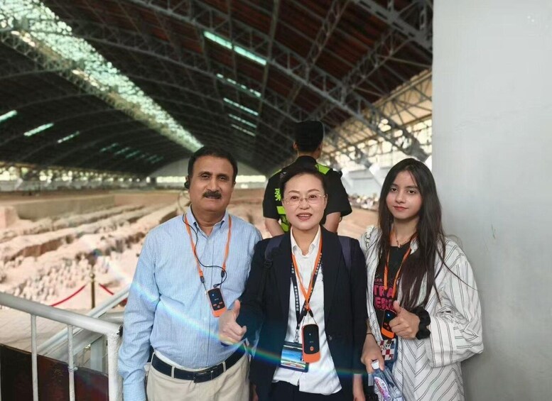 Three people standing in an open-air structure with a metal roof and a sandy ground. 5