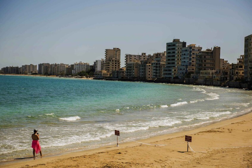 A beach with a person in a pink dress, a sandy shore, and a row of buildings in the background. 3