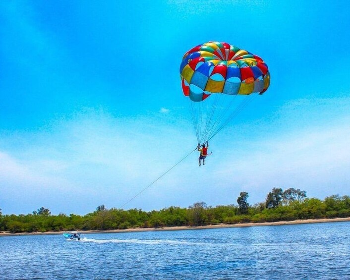 Iemand paraglide boven water, met een kleurrijke parachute, een jetski op de achtergrond en een kustlijn met begroeiing. 2