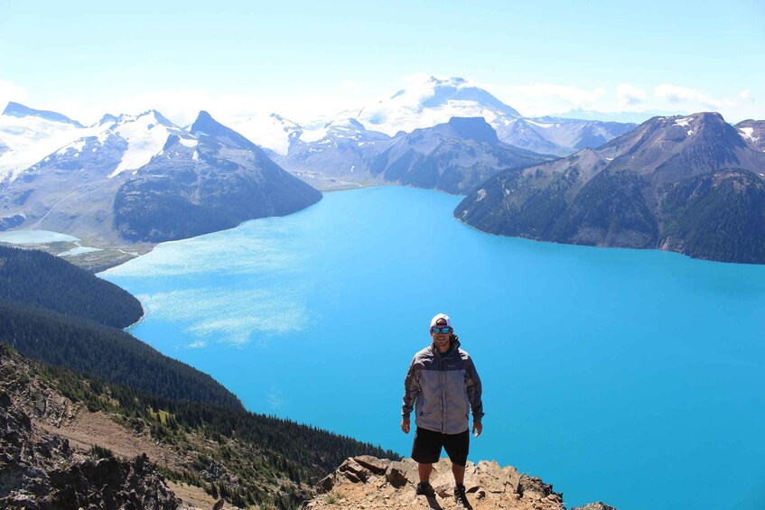 A person standing on a mountain ridge with a lake and snow-capped mountains in the background. 4