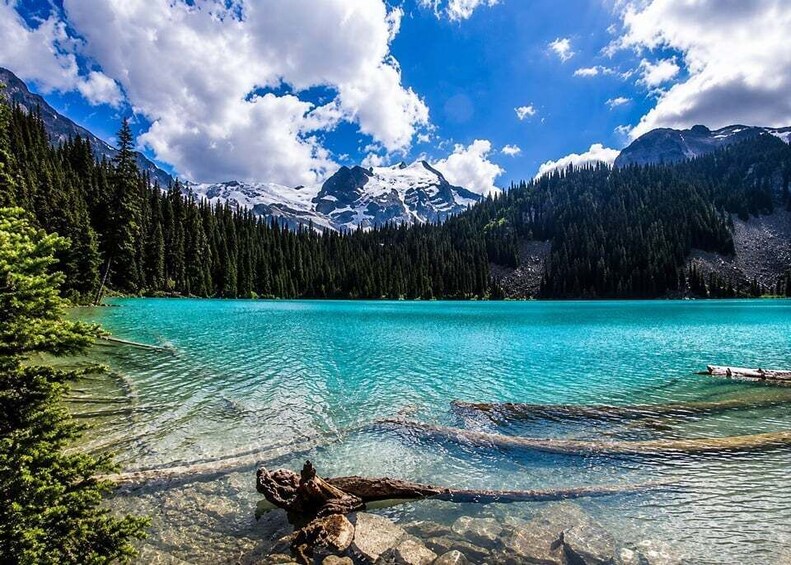 A clear turquoise lake surrounded by dense forest and snow-capped mountains under a partly cloudy sky. 5