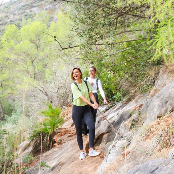 Two people hiking on a rocky trail surrounded by greenery. 1