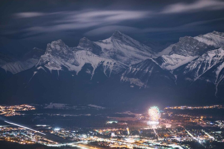 A snow-covered mountain range at night with a town below and a bright light in the distance. 2