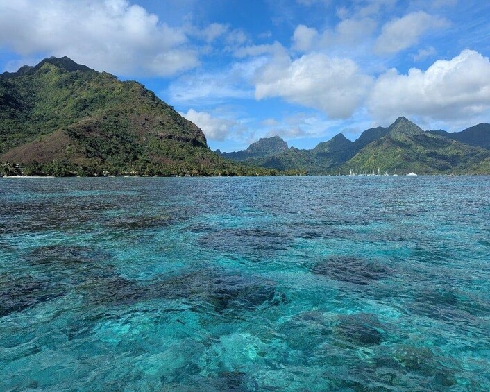 A clear turquoise sea with visible underwater rocks, a mountainous landscape, and a partly cloudy sky. 4