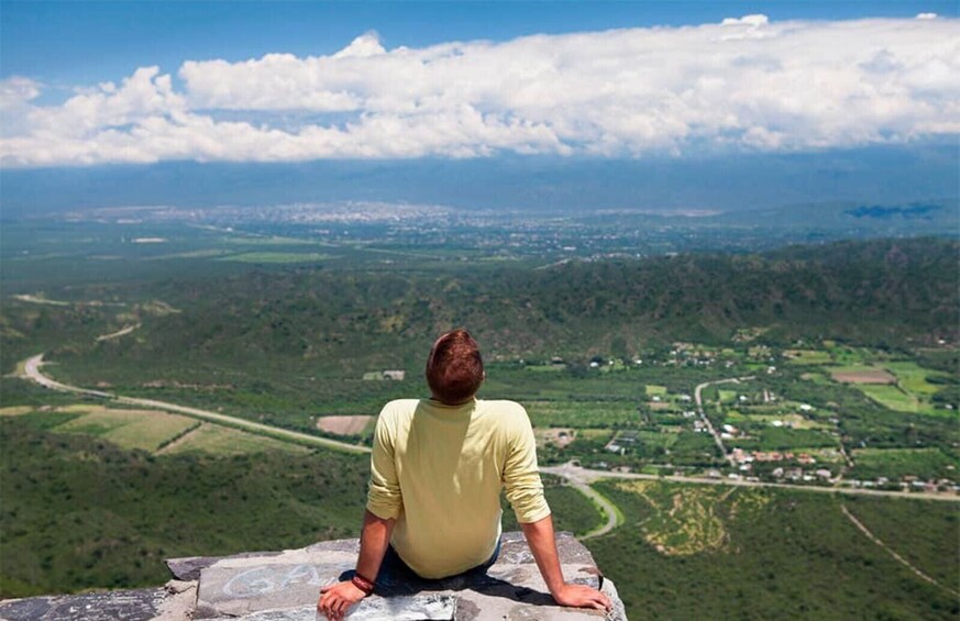 A person sitting on a rock overlooking a valley with fields and a distant town. 1