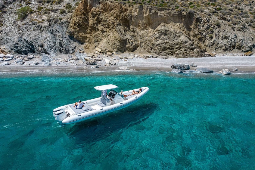 A white motorboat with two people on a clear turquoise sea, surrounded by rocky cliffs and a sandy beach. 2