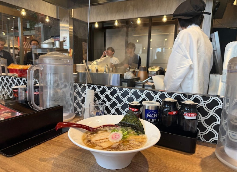 A bowl of ramen with a red chopstick, a clear mug, and condiments on a table in a restaurant. 3