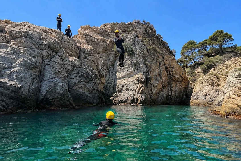 A person in a wetsuit and yellow helmet swimming in clear water, with rocky cliffs and trees in the background. 1