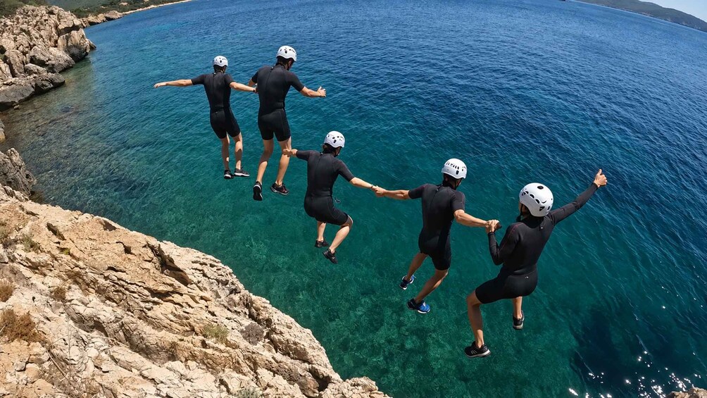 A group of people in wetsuits and helmets jumping off a rocky cliff into clear blue water. 3