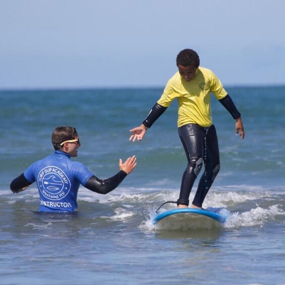 A person in a blue wetsuit with a logo, standing in the water, and another person in a yellow shirt riding a blue surfboard. 5