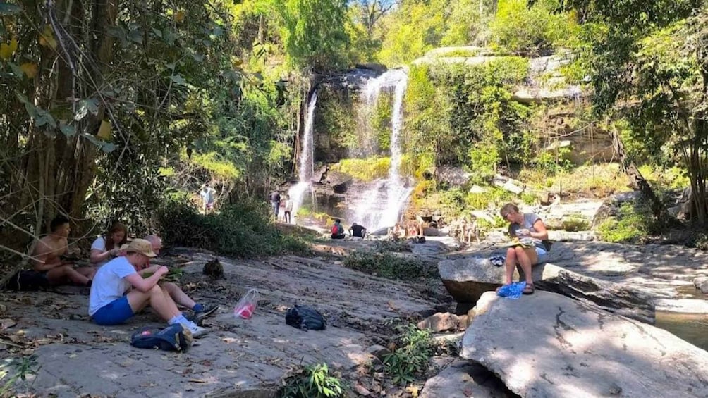 A waterfall surrounded by lush greenery, with people sitting and relaxing on rocks. 1