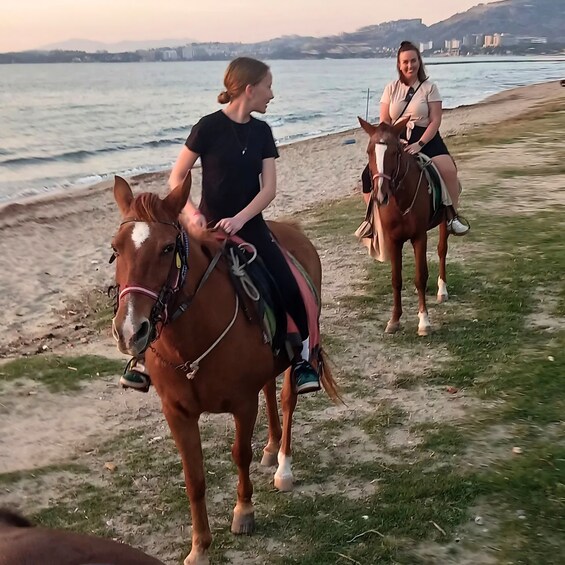Two people riding horses along a beach with a body of water and distant buildings in the background. 2