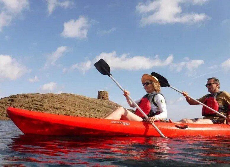 Two people kayaking in a red kayak on a calm body of water with a sandy shore and a small hill in the background. 4