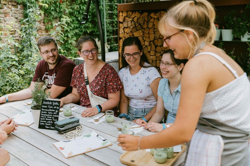 A group of people sitting at an outdoor table with a chalkboard menu, surrounded by greenery and a brick wall. 2