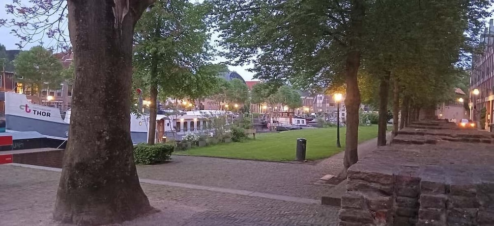A tree-lined pathway leading to a canal with boats, a bridge, and buildings in the background. 1