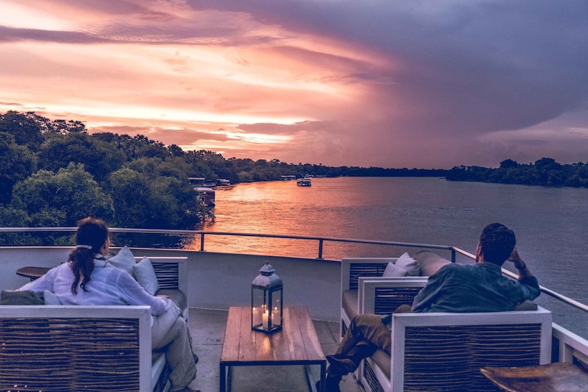 A sunset over a river with boats, a person sitting on a chair, and another person on a balcony. 2