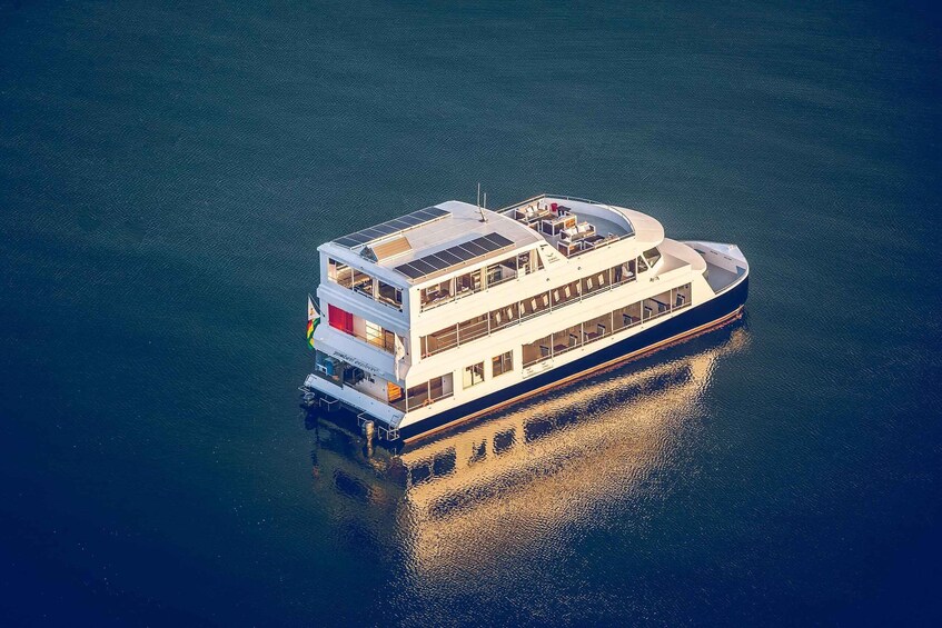 A large, multi-deck riverboat with a flat roof and solar panels, floating on calm water. 3