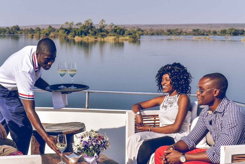 A waiter serving wine to a couple on a boat with a lake and trees in the background. 4