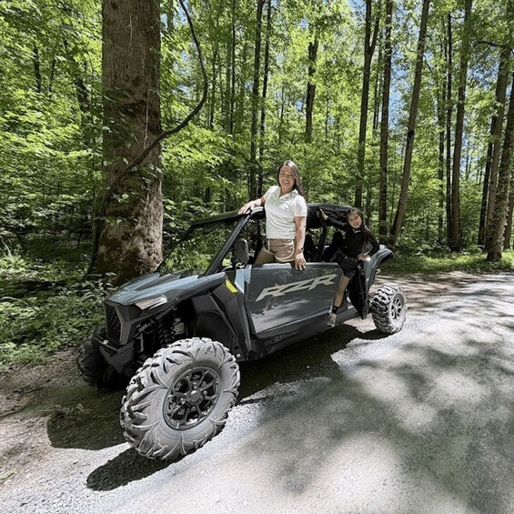 A person in a white shirt and khaki shorts sitting in a black ATV with large tires on a forest path. 1