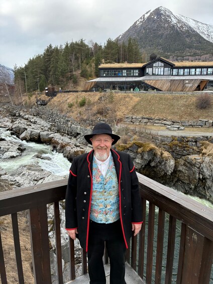 A man standing on a wooden bridge with a river and mountains in the background. 1