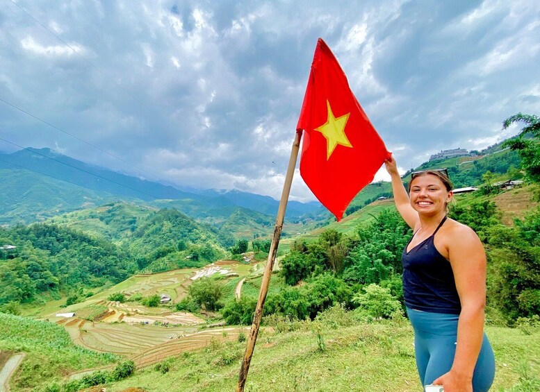 A person holding a flag with a star, standing in a hilly landscape with terraced fields. 1