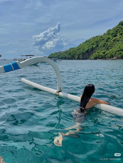 A person swimming in clear blue water with a boat and a forested hill in the background. 1