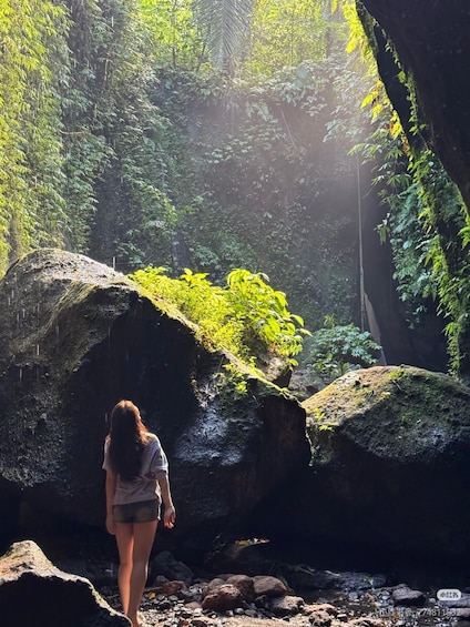 A person standing in a forested area with large rocks and sunlight filtering through the trees. 2